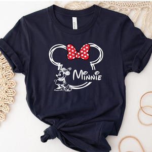 May include: A navy blue t-shirt with a white outline of Minnie Mouse's head and ears, a red bow with white polka dots, and the text "Minnie" underneath.