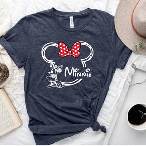 May include: A dark blue t-shirt with a white outline of Minnie Mouse's head and ears. The ears have a red bow with white polka dots. The text "Minnie" is written below the ears.