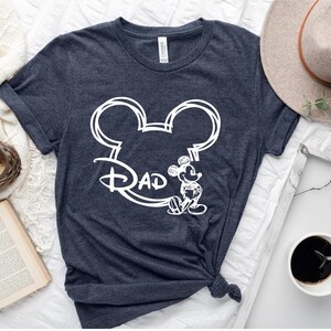 Pu&ograve; includere: Una maglietta blu scuro con un disegno bianco a forma di testa di Topolino e la parola "Pap&agrave;" all'interno delle orecchie.