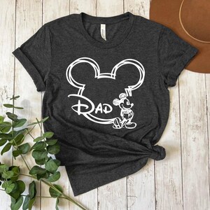 Pu&ograve; includere: Una maglietta grigia scura con un contorno bianco della testa di Topolino e la parola "Dad" all'interno delle orecchie. Topolino &egrave; in piedi a destra della testa.