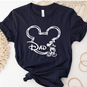 Pu&ograve; includere: Una maglietta blu navy con un contorno bianco della testa di Topolino e la parola "Dad" all'interno delle orecchie. Topolino &egrave; in piedi a destra della testa.