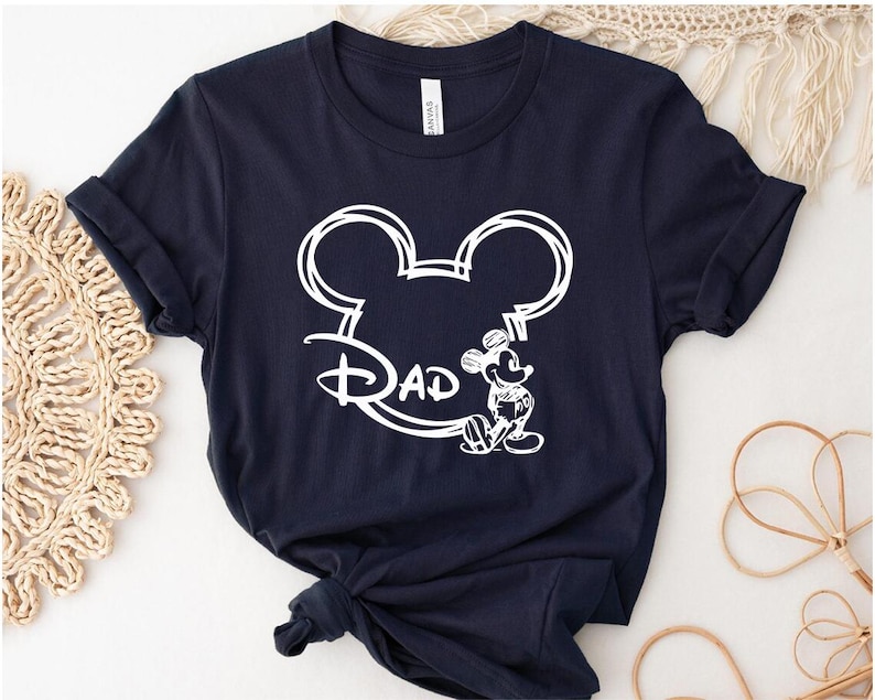 Pu&ograve; includere: Una maglietta blu navy con un contorno bianco della testa di Topolino e la parola "Dad" all'interno delle orecchie. Topolino &egrave; in piedi a destra della testa.