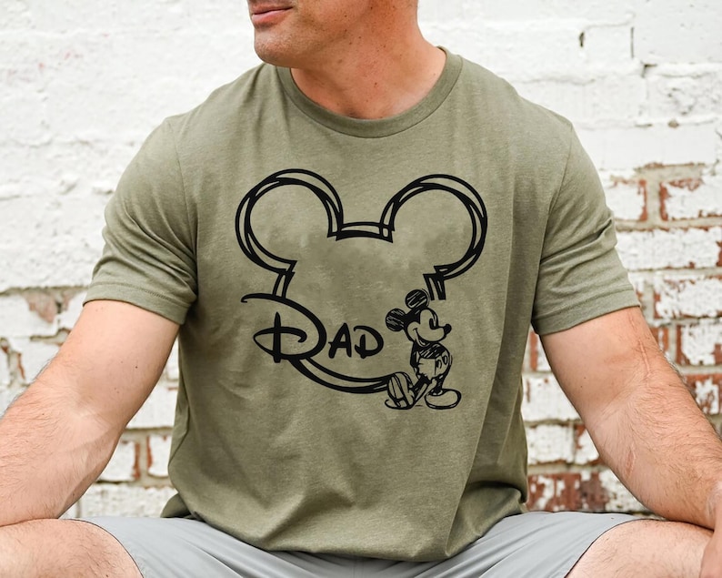 Pu&ograve; includere: Una maglietta verde con un contorno nero della testa di Topolino e la parola "Dad" all'interno delle orecchie. Topolino &egrave; in piedi a destra della testa.