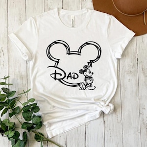 Pu&ograve; includere: Maglietta bianca con un contorno nero della testa di Topolino e la parola "Dad" all'interno. Topolino &egrave; in piedi a destra della testa.