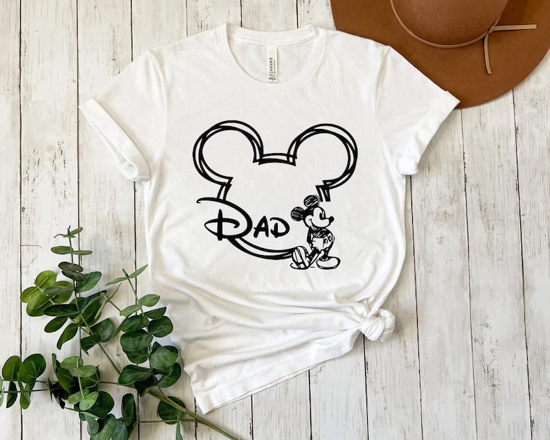 Pu&ograve; includere: Maglietta bianca con un contorno nero della testa di Topolino e la parola "Dad" all'interno. Topolino &egrave; in piedi a destra della testa.