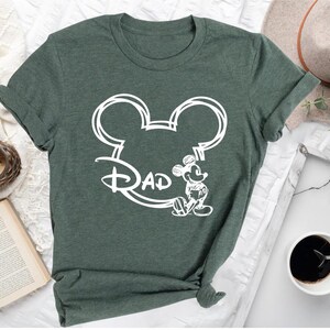 Pu&ograve; includere: Una maglietta verde con un contorno bianco della testa di Topolino e la parola "DAD" all'interno delle orecchie. Topolino &egrave; in piedi a destra della testa.