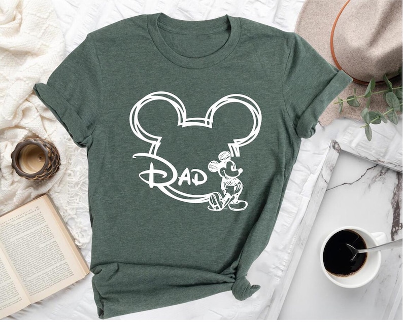 Pu&ograve; includere: Una maglietta verde con un contorno bianco della testa di Topolino e la parola "DAD" all'interno delle orecchie. Topolino &egrave; in piedi a destra della testa.