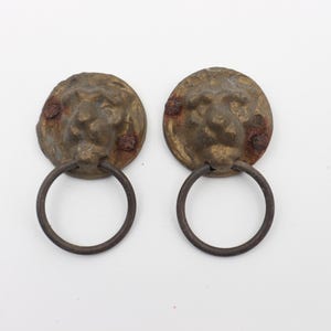 Pode incluir: Dois puxadores de porta antigos de latão com forma de cabeça de leão e alças de anel. As cabeças de leão têm um acabamento desgastado e os anéis são de metal escuro.