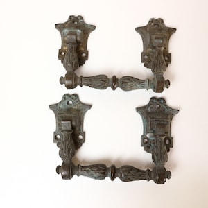 Könnte beinhalten: Vier antike Türgriffe aus Bronze. Jeder Griff hat ein verziertes Design mit einer dekorativen Rückplatte und einem gebogenen Griff. Die Bronze hat eine verwitterte Patina mit grünen und braunen Farbtönen.
