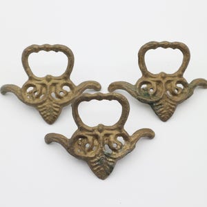 Könnte beinhalten: Drei goldfarbene, verzierte Metall-Dekorationsteile. Jedes Teil hat einen Schlaufe-Griff und eine spitze Unterseite.