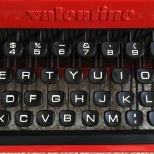Peut inclure: Une machine à écrire rouge avec des touches noires et le mot "valentino" imprimé en haut. Les touches ont des lettres, des chiffres et des symboles.