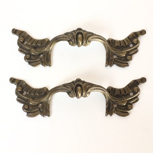 Könnte beinhalten: Zwei verzierte, goldfarbene Metall-Schubladengriffe mit einem dekorativen, floralen Design. Die Griffe haben eine geschwungene Form und sind für die Anbringung an Möbeln konzipiert.