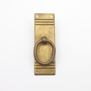 Peut inclure: Une poignée de porte en laiton avec une poignée en forme d'anneau. La poignée est montée sur une plaque rectangulaire avec des rainures décoratives.
