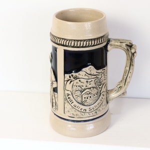May include: A ceramic beer stein with a blue and white design. The stein features a scene of a mountain village with the text "Heit allen Schmarnz" on the front.
