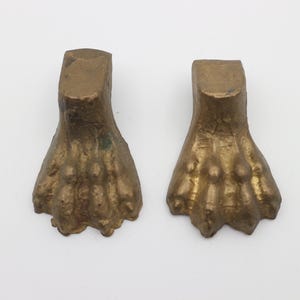 Op de afbeelding: Twee goudkleurige metalen meubelpoten in de vorm van een leeuwenpoot met vier tenen. De poten zijn licht beschadigd en hebben een getextureerd oppervlak.