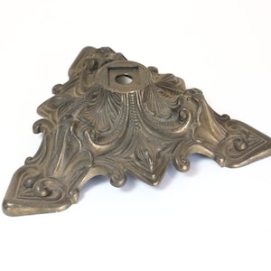 May include: A gold-toned metal base with an ornate, intricate design. The base has a round top with a hole in the center and three legs that extend out in a triangular shape.