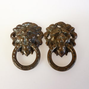 Pode incluir: Duas aldravas antigas de bronze com cabeça de leão. Cada aldrava apresenta um rosto de leão detalhado e uma pega em forma de anel. O bronze tem uma pátina desgastada, adicionando à estética vintage.