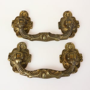 Könnte beinhalten: Zwei verzierte Messing-Schubladengriffe mit einem dekorativen, gerollten Design. Die Griffe haben eine quadratische Öffnung in der Mitte zum Befestigen.