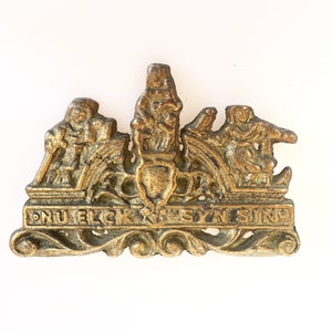 Könnte beinhalten: Eine goldfarbene Metallstifte mit einem Design von drei Figuren in einem Wagen, mit dem Text "DNU ELCK SYN SING" darunter.
