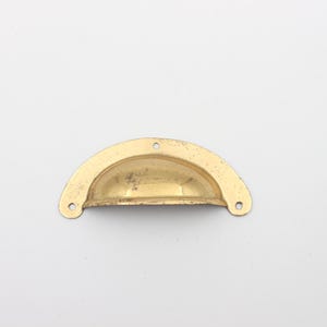 Op de afbeelding: Een gouden metalen kastknop met een afgeronde, halve cirkelvorm. De knop heeft twee kleine gaten voor montage.