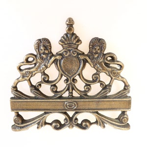 Op de afbeelding: Een decoratieve metalen plaquette in goudkleur met een kroon en twee leeuwen aan weerszijden. De plaquette heeft een rechthoekige ruimte voor tekst.