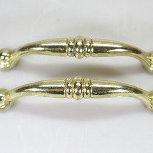 Könnte beinhalten: Zwei goldfarbene Schrankgriffe mit abgerundeter, leicht gebogener Form und einem dekorativen, erhabenen, mittigen Band.