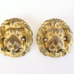 Könnte beinhalten: Zwei antike, goldfarbene Löwenkopf-Schrankgriffe. Jeder Griff zeigt ein detailliertes Löwengesicht mit strukturierter Mähne, Nase und offenem Maul. Die Griffe haben zwei kleine Löcher zur Befestigung und sind wahrscheinlich aus Gussmetall.