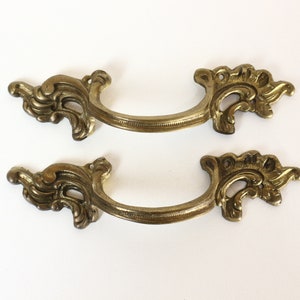 Könnte beinhalten: Zwei verzierte, goldfarbene Metall-Schubladengriffe mit einem dekorativen Schnörkeldesign.