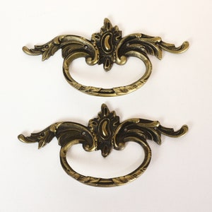 Könnte beinhalten: Zwei verzierte, goldfarbene Metall-Schubladengriffe mit einem dekorativen Schnörkeldesign.