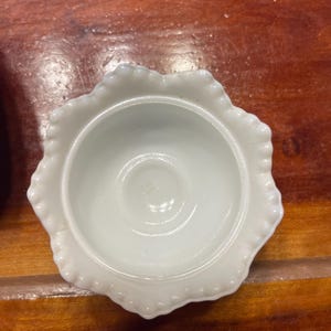 May include: A small, white, scalloped-edge dish with a central indentation. The dish has a glossy finish and is set against a warm, wooden surface. The dish's rim features a delicate, beaded design.