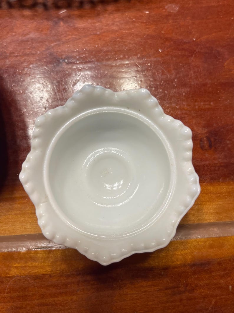 May include: A small, white, scalloped-edge dish with a central indentation. The dish has a glossy finish and is set against a warm, wooden surface. The dish's rim features a delicate, beaded design.