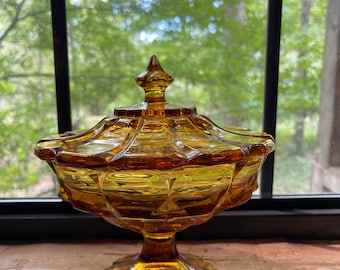May include: A vintage amber glass candy dish with a fluted design and a domed lid. The dish sits on a pedestal base and has a delicate, ornate design.