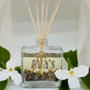 May include: A clear glass diffuser bottle with a label that reads "Ava's". The bottle is filled with a light brown liquid and has brown stones at the bottom.  The bottle has wooden diffuser sticks sticking out of the top.