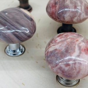 May include: Four decorative cabinet knobs with a marbled design. The knobs are made of a light pink and gray stone with a glossy finish. The knobs have a round shape and are attached to a metal base.