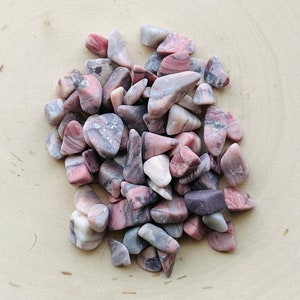 May include: A collection of tumbled stones featuring a mix of dusty pink, mauve, and grey tones. Each stone has a smooth, polished surface with natural veining and irregular shapes, creating a varied texture across the entire cluster of minerals.