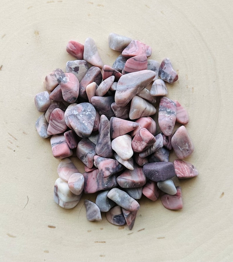 May include: A collection of tumbled stones featuring a mix of dusty pink, mauve, and grey tones. Each stone has a smooth, polished surface with natural veining and irregular shapes, creating a varied texture across the entire cluster of minerals.