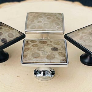 May include: Four square cabinet knobs featuring a polished stone inlay with a circular pattern. Two knobs have matte black metal bases and frames, while the other two have polished silver-tone metal bases and frames, arranged in a group.