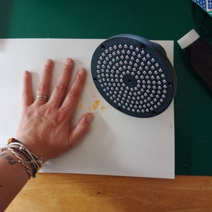May include: A white surface with a black circular tool with silver studs. The tool is surrounded by a yellow stain. A hand with a ring and bracelets is resting on the surface.