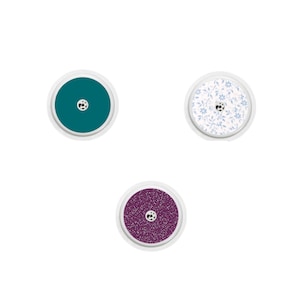 May include: Three round adhesive stickers with different designs. The first sticker is teal blue, the second is white with a blue floral pattern, and the third is a sparkly purple.