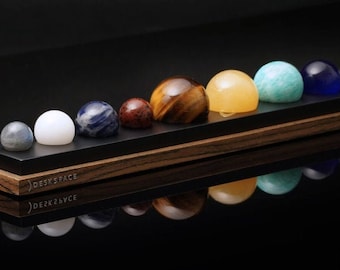 Puede incluir: Un conjunto de nueve esferas de piedras preciosas que representan los planetas, dispuestas sobre un expositor negro y con vetas de madera. Las esferas varían en color, incluyendo blanco, azul, marrón, amarillo y verde. El expositor tiene el texto "DESKSPACE".