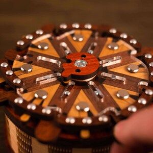Puede incluir: Una caja de rompecabezas de madera con una tapa circular y una perilla de madera roja. La caja est&aacute; hecha de diferentes tipos de madera, incluyendo nogal y arce. La parte superior de la caja est&aacute; decorada con un patr&oacute;n geom&eacute;trico.