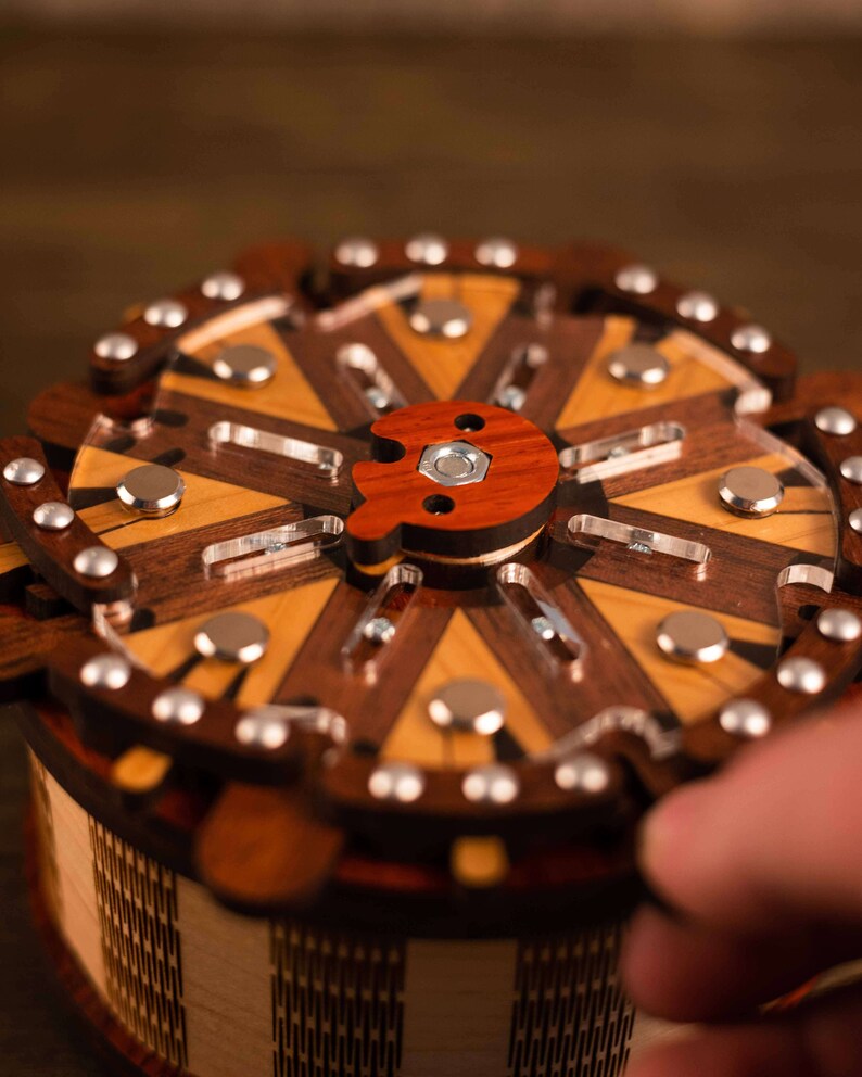 Puede incluir: Una caja de rompecabezas de madera con una tapa circular y una perilla de madera roja. La caja est&aacute; hecha de diferentes tipos de madera, incluyendo nogal y arce. La parte superior de la caja est&aacute; decorada con un patr&oacute;n geom&eacute;trico.