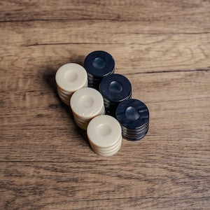 Puede incluir: Un conjunto de fichas de damas negras y blancas apiladas en una fila sobre una superficie de madera. Las fichas est&aacute;n dispuestas en colores alternos, con las fichas negras encima de las fichas blancas.