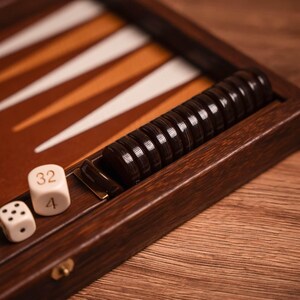 Puede incluir: Primer plano de un juego de backgammon de madera. El tablero presenta triángulos marrones, blancos y naranjas. Las fichas de juego de color marrón oscuro se guardan en un compartimento lateral. Se ven dos dados con los números 4 y 32.