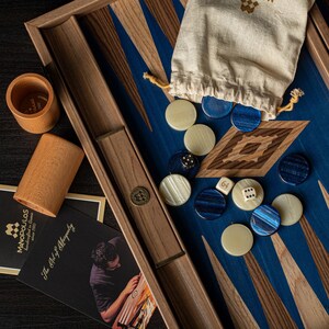 Könnte beinhalten: Ein Backgammon-Brett aus Holz mit blauen und braunen Einlegearbeiten. Das Brett verfügt über ein eingebautes Fach zur Aufbewahrung der Spielsteine. Die Spielsteine sind weiß und blau und liegen verstreut auf dem Brett. Außerdem sind zwei Würfel und ein kleiner Beutel zu sehen.