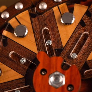 Puede incluir: Primer plano de un rompecabezas de madera con una pieza central en forma de oso. El rompecabezas tiene m&uacute;ltiples capas de madera con diferentes colores y patrones. El rompecabezas tiene una variedad de detalles met&aacute;licos, incluidos tornillos y peque&ntilde;as piezas redondas.