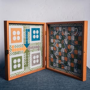 May include: Wooden board game featuring two classic games: Snakes and Ladders and a colorful board game with arrows and squares. The game is housed in a wooden box with a brass clasp. The board games have orange, green, blue, and white colors.