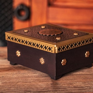 May include: A black wooden box with a gold trim and decorative metal accents. The top of the box has a circular design with a raised center.