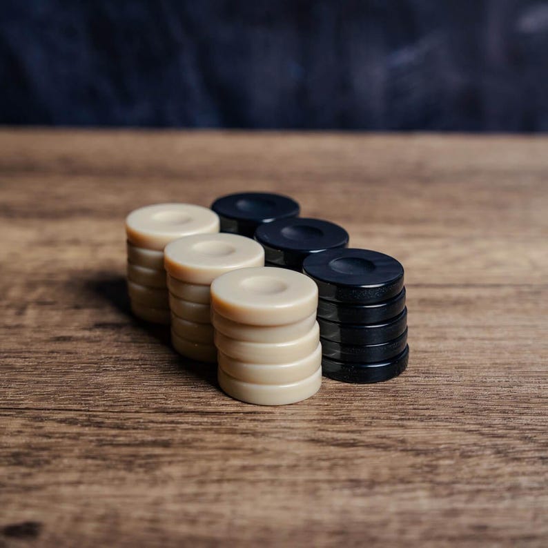 Puede incluir: Un conjunto de damas negras y blancas apiladas en una fila sobre una superficie de madera. Las damas est&aacute;n dispuestas en colores alternos, con las damas negras a la derecha y las damas blancas a la izquierda.