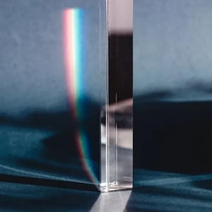 Può includere: Un prisma rettangolare trasparente rifrange la luce, creando un vibrante spettro arcobaleno su una superficie blu scuro. I bordi del prisma sono ben definiti, riflettendo la luce. L'effetto complessivo è un gioco di luce e colore.